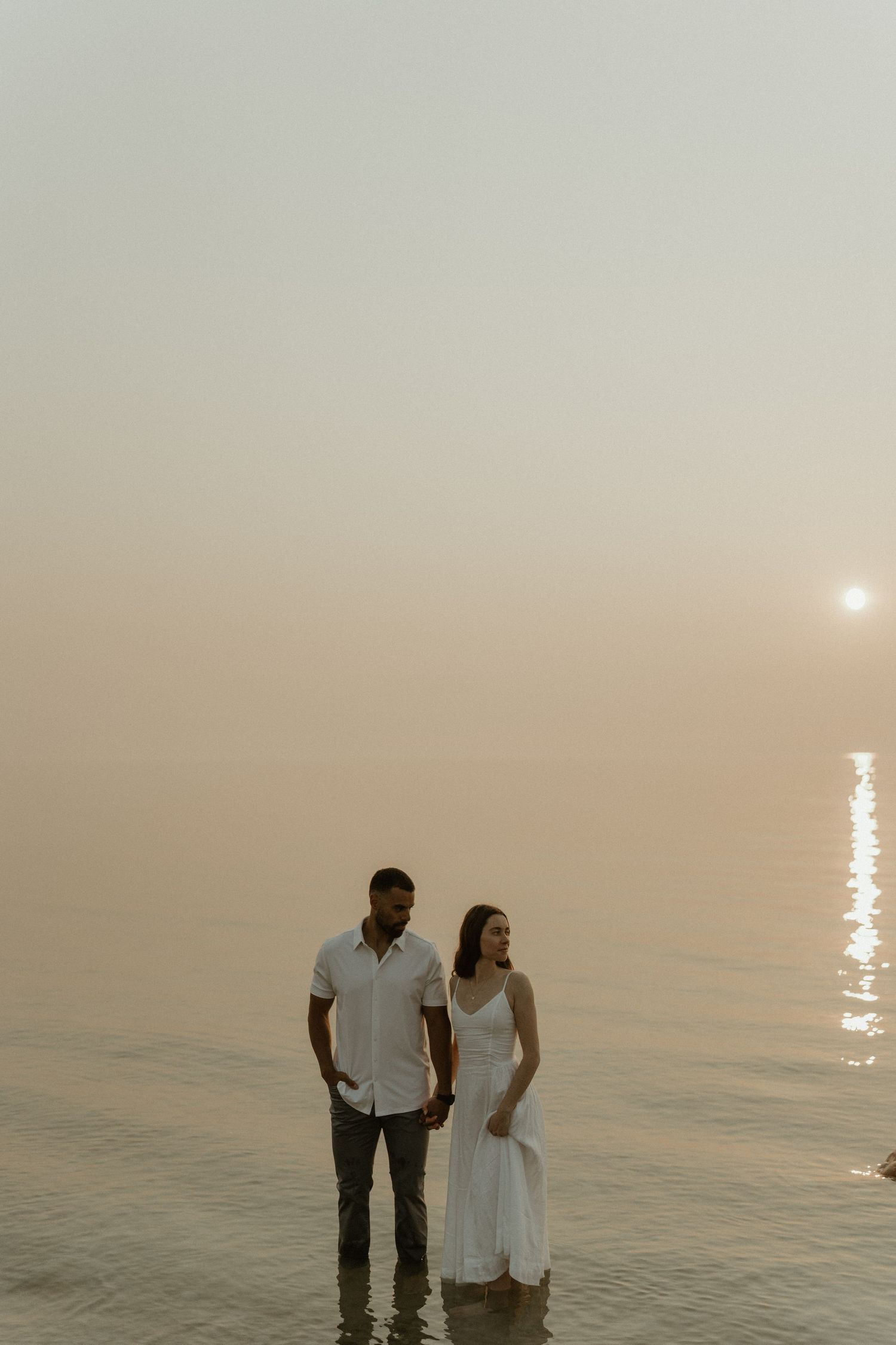 A couple stands together on a beach at sunset with the warm glow of the sun reflecting on the calm ocean waters.
