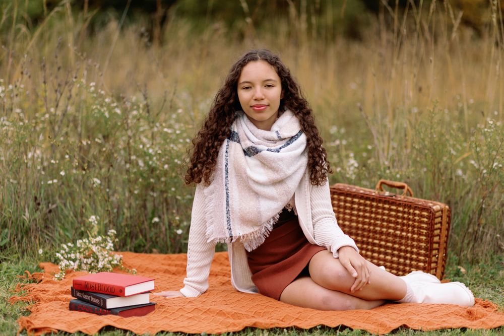 Person sitting on orange blanket in autumn field with books and wicker basket enjoying outdoor leisure time.