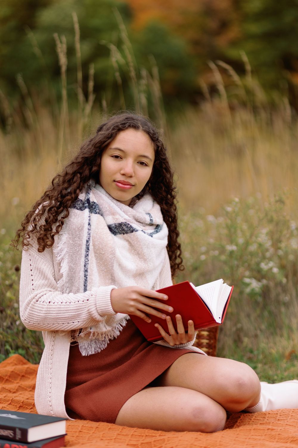 Person reading a red book while sitting on an orange blanket outdoors in a field with tall grass on a fall day.