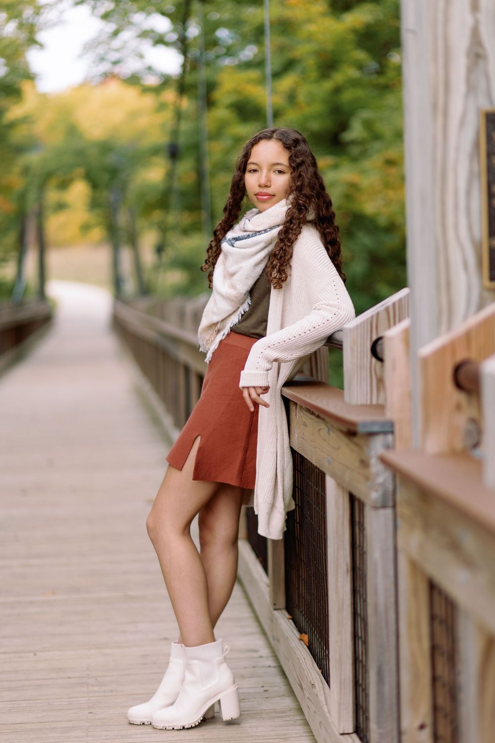 Stylish autumn outfit with white sweater, terracotta skirt and boots showcased on wooden bridge surrounded by fall foliage.