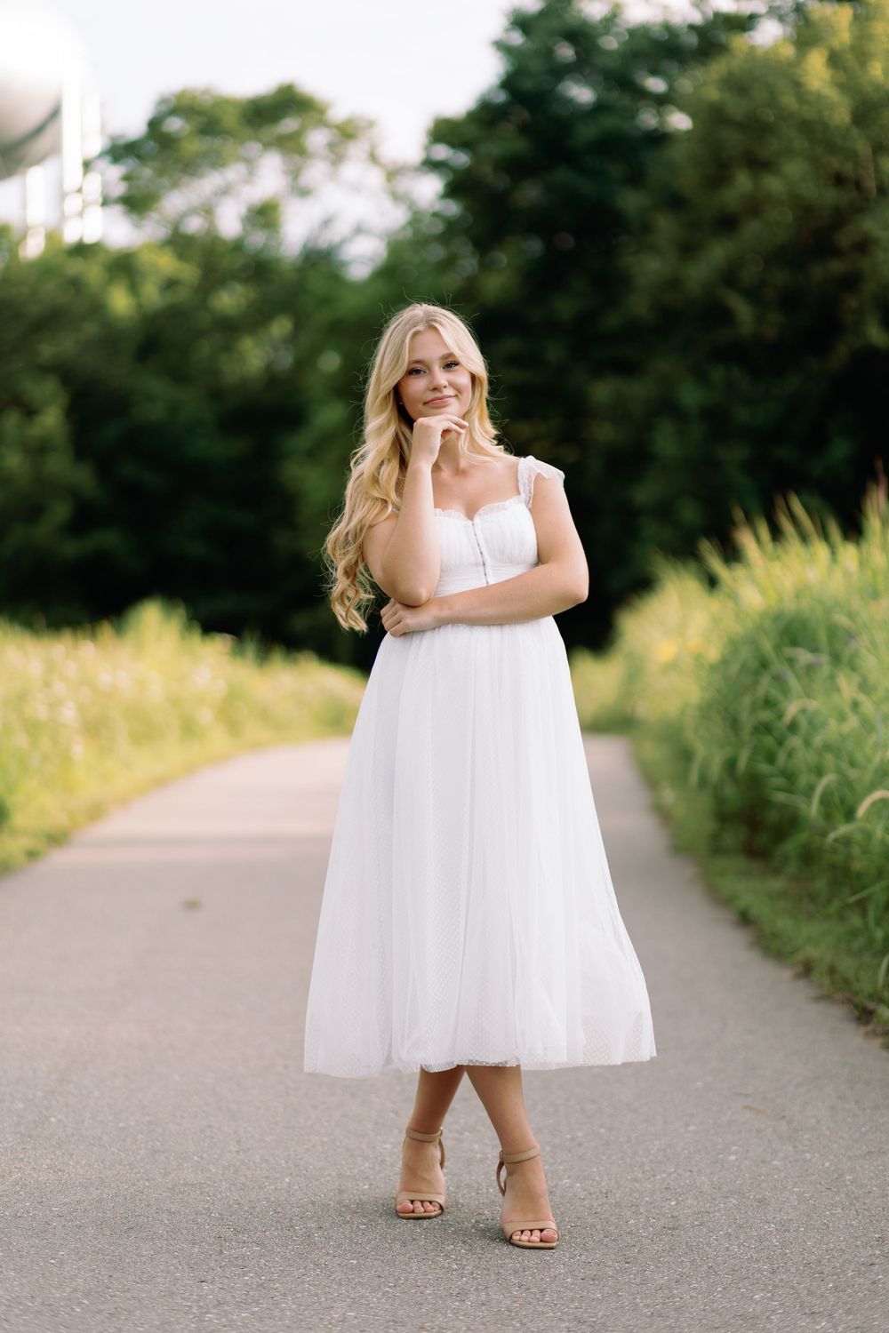 A woman in a white midi-length dress and sandals poses outdoors on a paved path surrounded by green foliage.