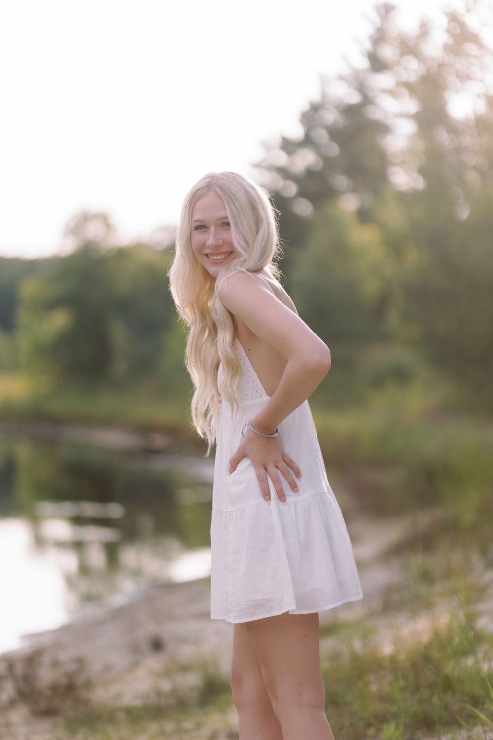 A person wearing a white summer dress poses by a lakeside during golden hour lighting.