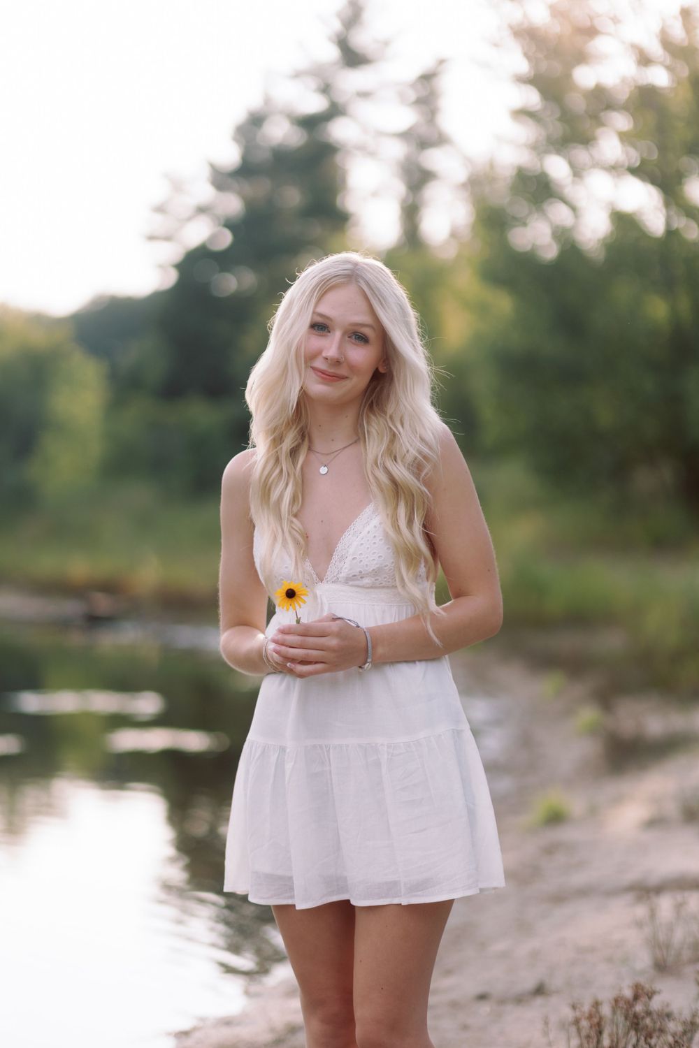 A person in a white summer dress holds a sunflower while standing near a peaceful lakeside at sunset.