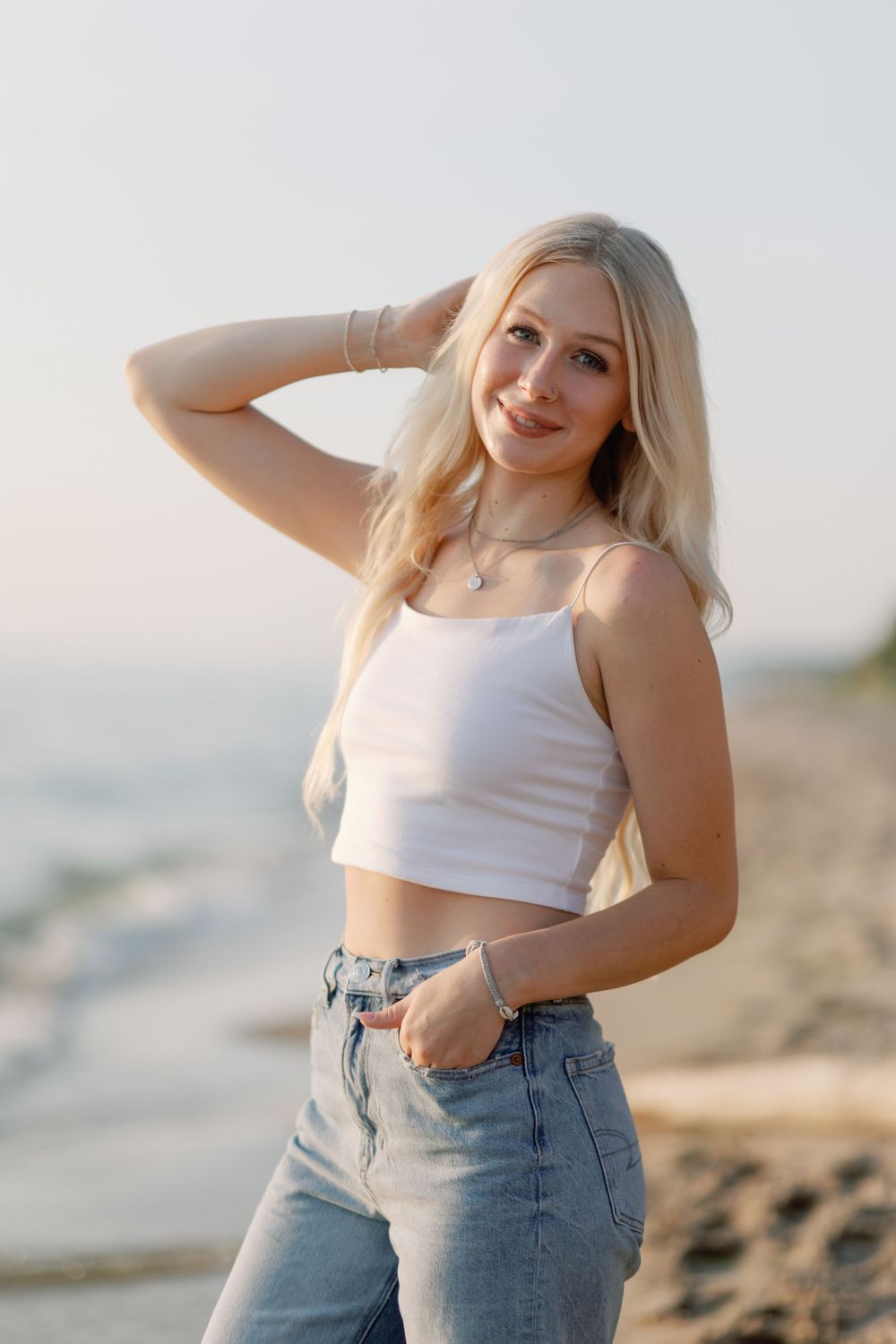 High school senior in white crop top and jeans poses against coastal sunset backdrop, holland beach, grand rapids.