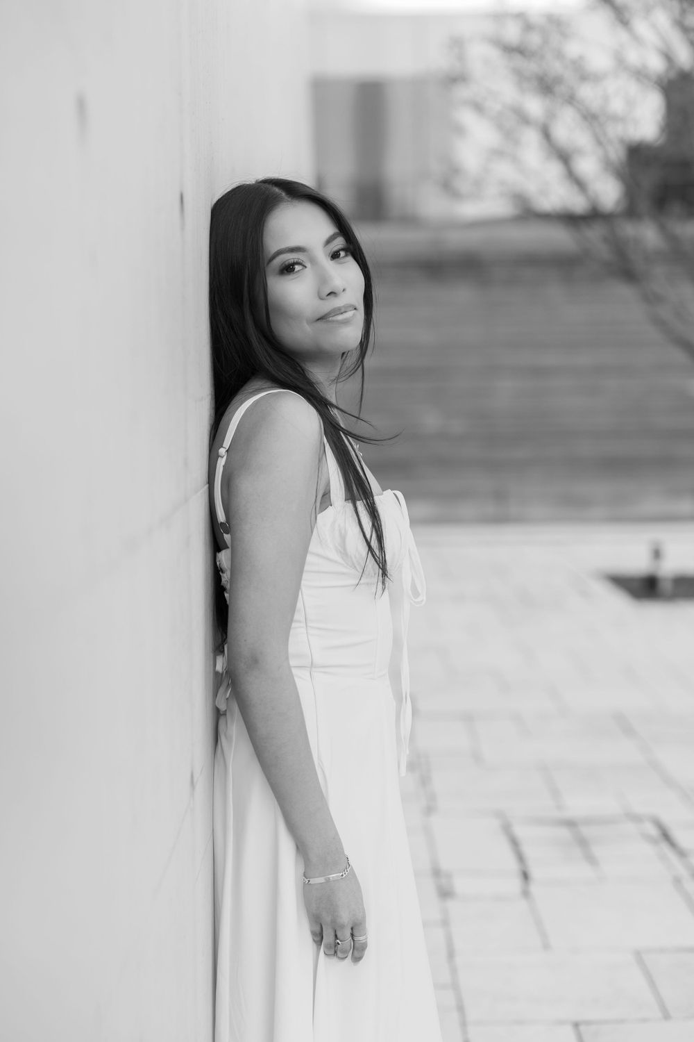Black and white portrait of a person in a white dress leaning against a wall outdoors.