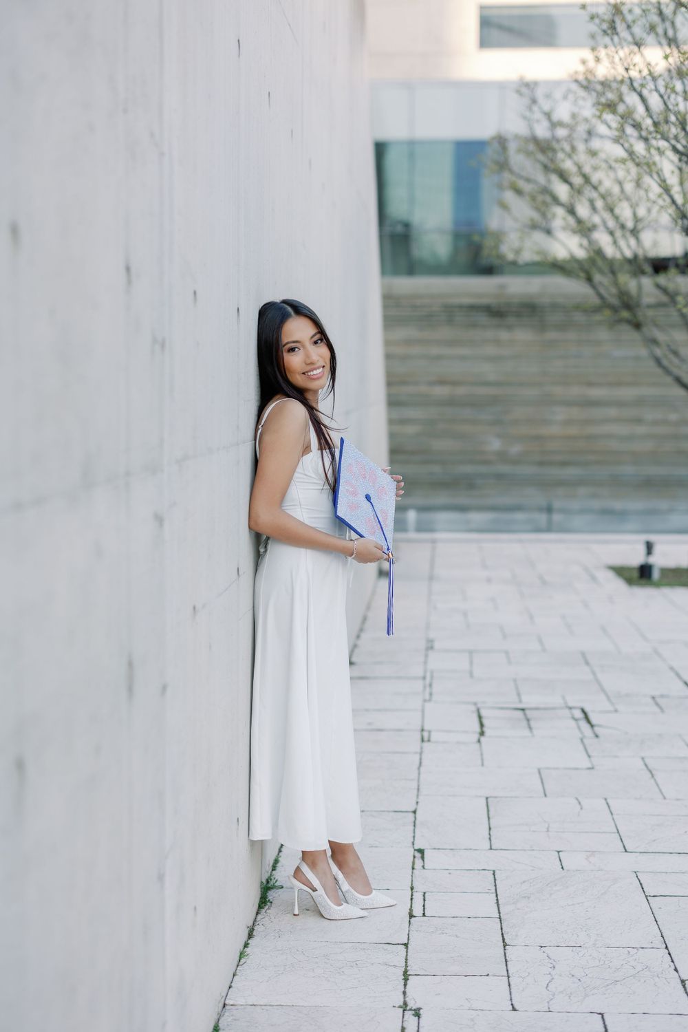 A person in a white sleeveless dress and heels poses against a concrete wall on a stone paved walkway.