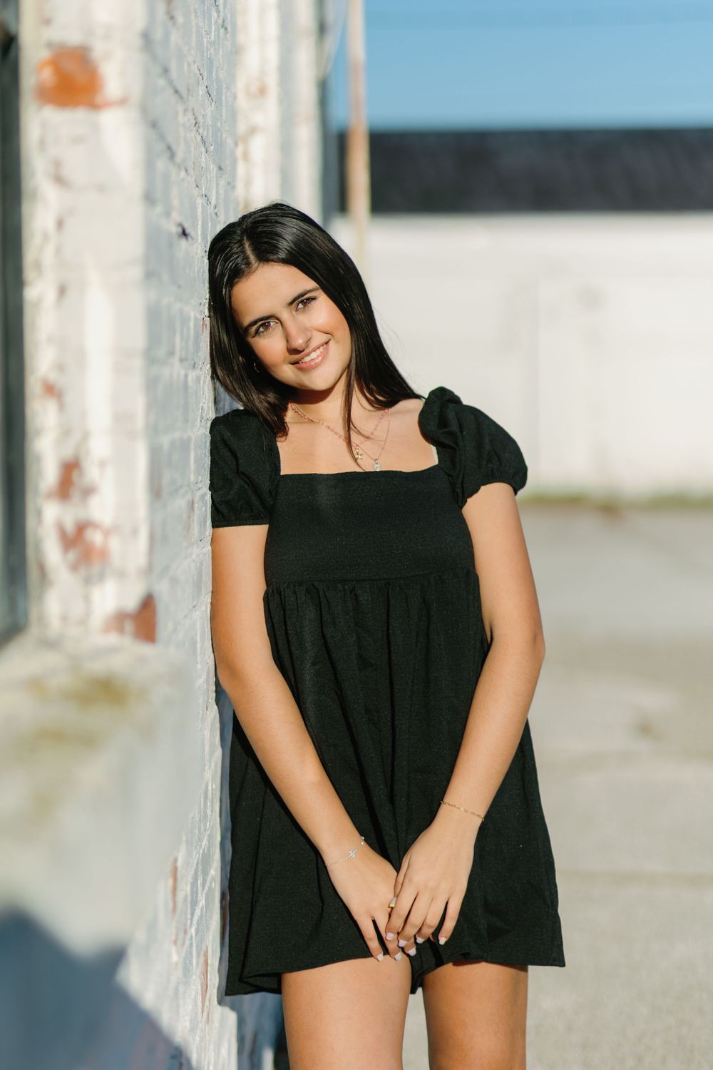 High school senior in a black babydoll dress poses casually against a light colored wall on a sunny day, senior photos, grand rapids.