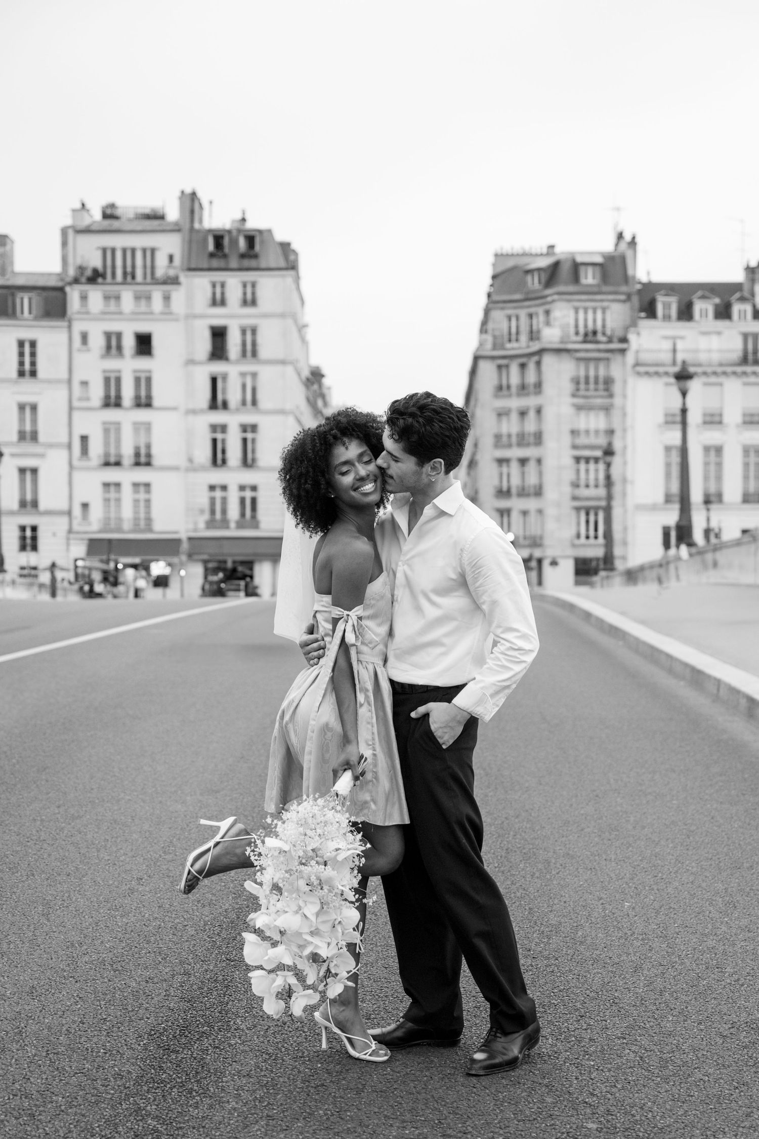 Couple in wedding attire sharing a joyful moment on a charming Parisian street with cafe lights at dusk.