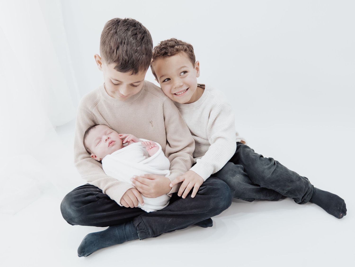 A young child in sage green overalls and white t-shirt sits barefoot on a white background. Day nursery photoshoot