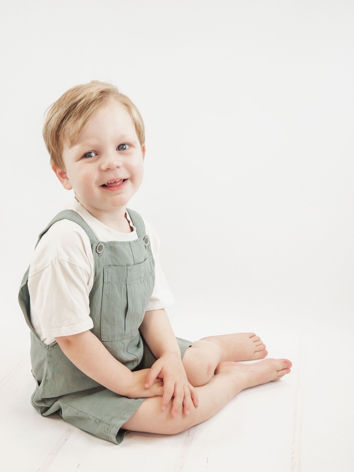 A young child in sage green overalls and white t-shirt sits barefoot on a white background. Day nursery photoshoot