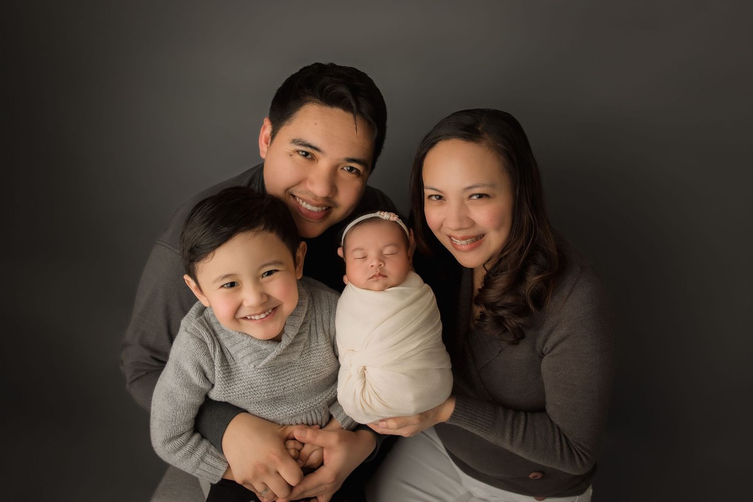 A couple holds their sleeping newborn baby while wearing a floral dress and grey shirt in a tender family portrait.