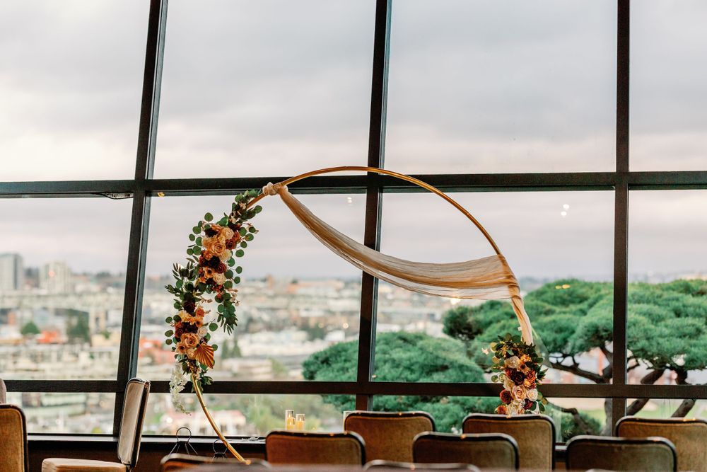 Circular floral wedding arch with orange blooms and ribbon overlooking city skyline through large windows.