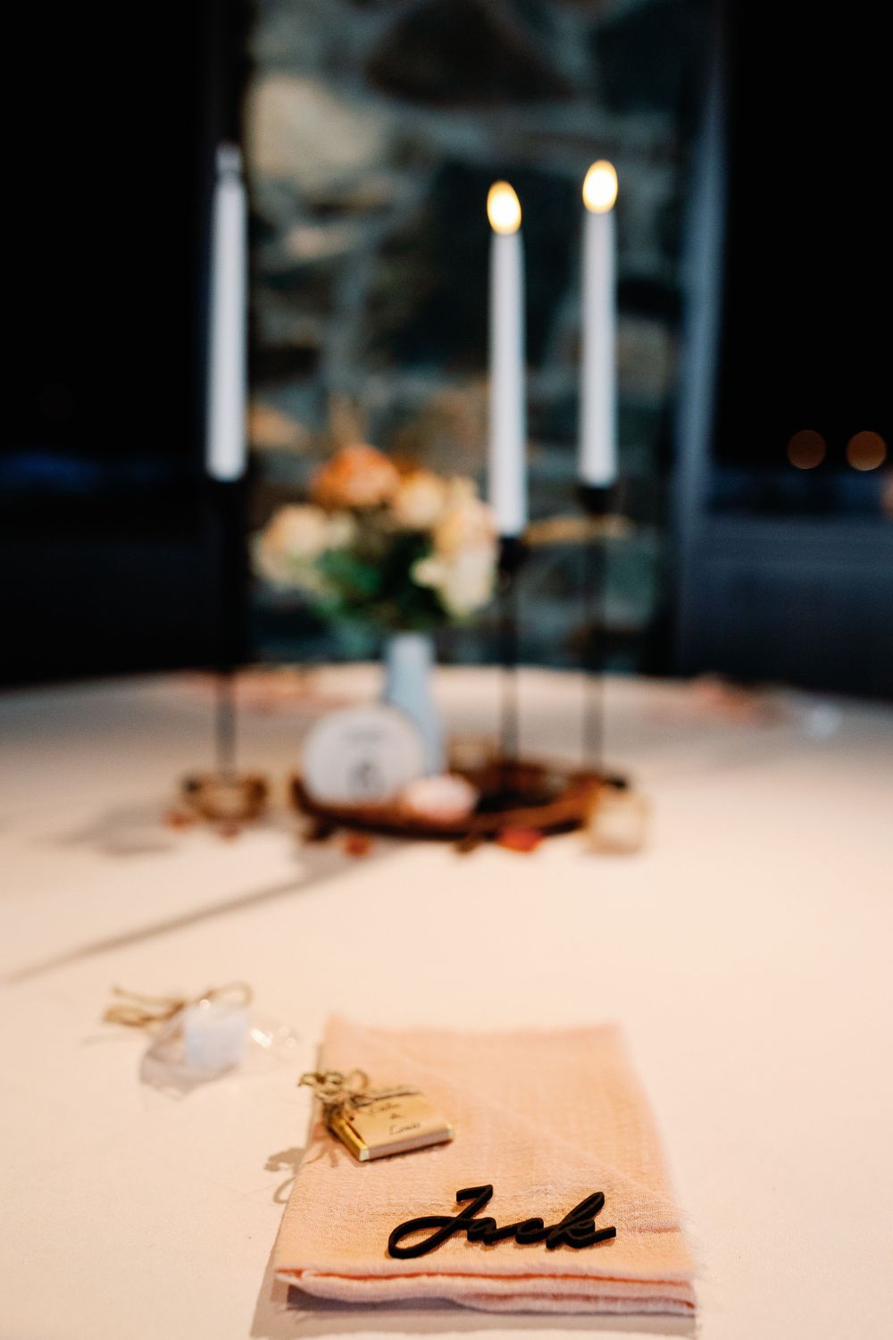 Wedding place setting with name card reading Josh on blush napkin with gold locket favor and candlelit centerpiece.