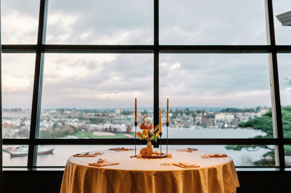 Intimate sweetheart table with gold tablecloth and tall candle centerpiece before panoramic city view windows at dusk.