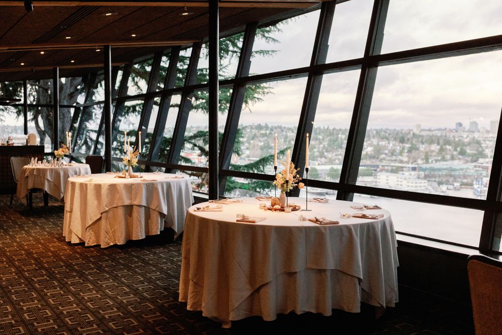 Elegant wedding reception room with white tablecloths and panoramic floor-to-ceiling windows overlooking a city skyline.