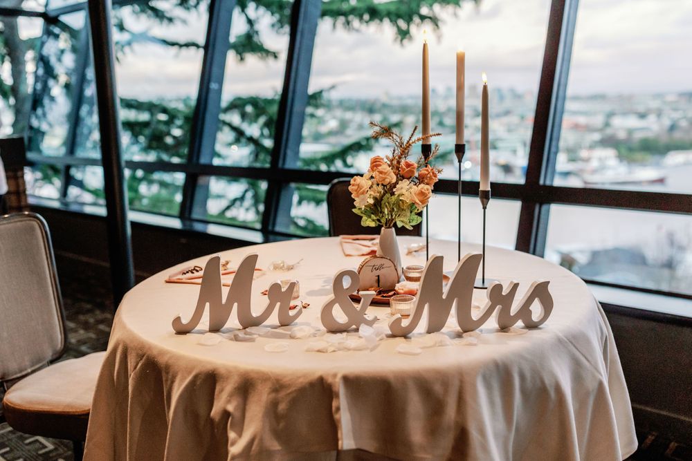 Wedding sweetheart table with Mr & Mrs wooden signs, candles, and floral centerpiece by large windows.