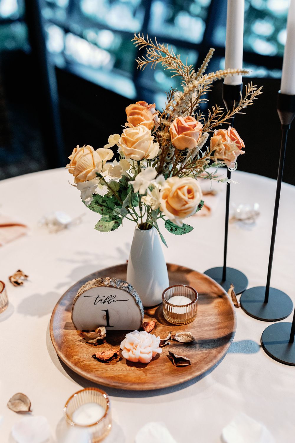 Wedding table centerpiece with peach roses in a vase, candles, and a wooden table number on a round tray.