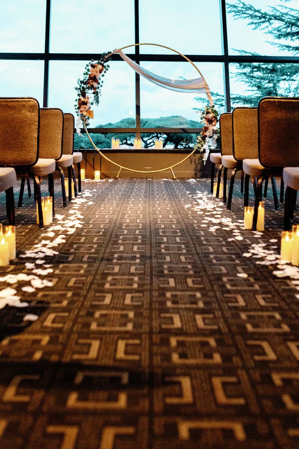 Wedding ceremony aisle with candles, rose petals, and a golden circular floral arch in front of panoramic windows.