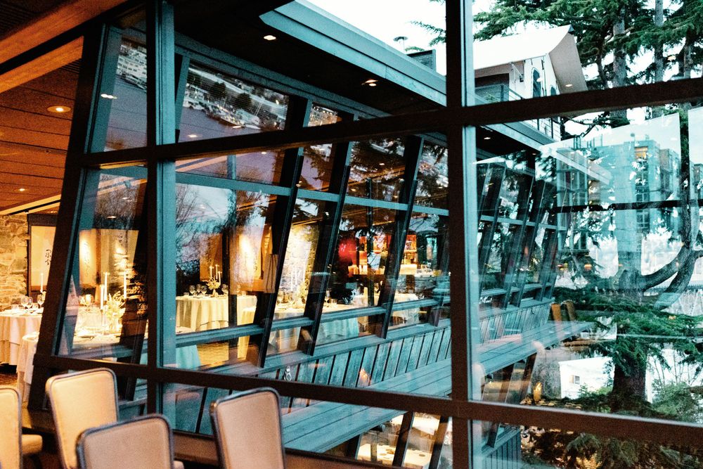 Modern wedding venue exterior with large glass windows reflecting elegant reception decor and lush trees.