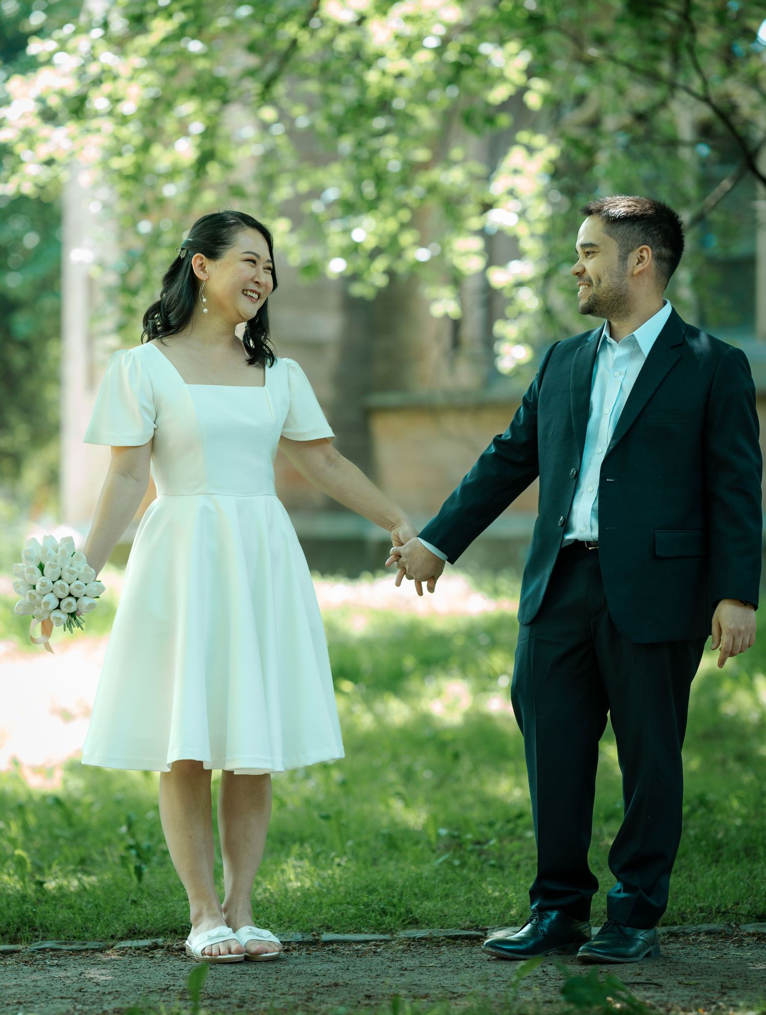 Couple in wedding attire holding hands while walking on garden path during sunny day.