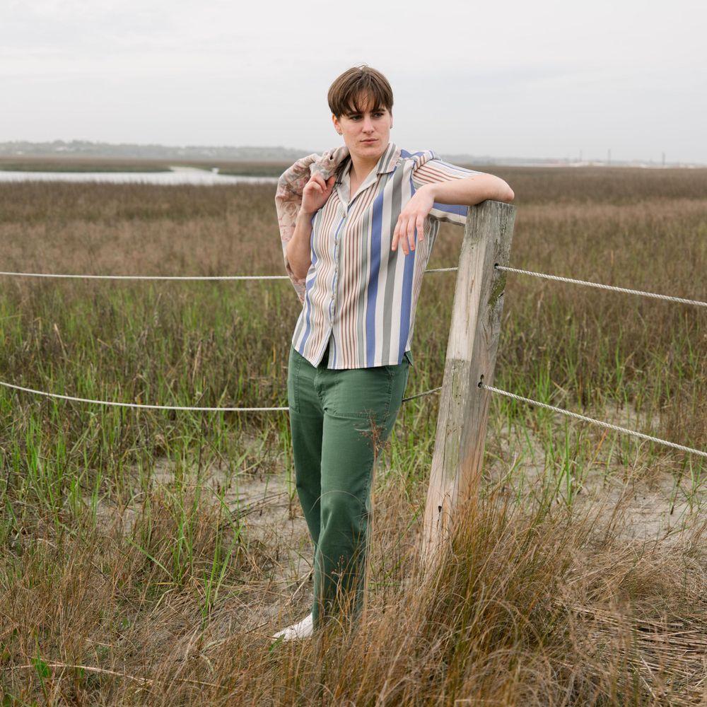 Casual pose leaning against a wooden fence post wearing striped shirt and green pants.