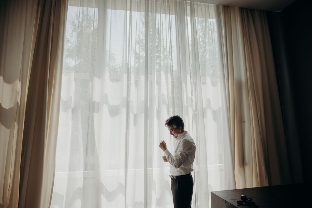 Man fixing his shirt and getting ready on his wedding day by a wide, bright window