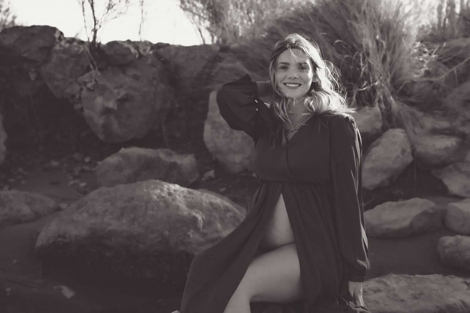 Black and white artistic portrait showing a person in dark clothing posed among large rocks in a natural outdoor setting.