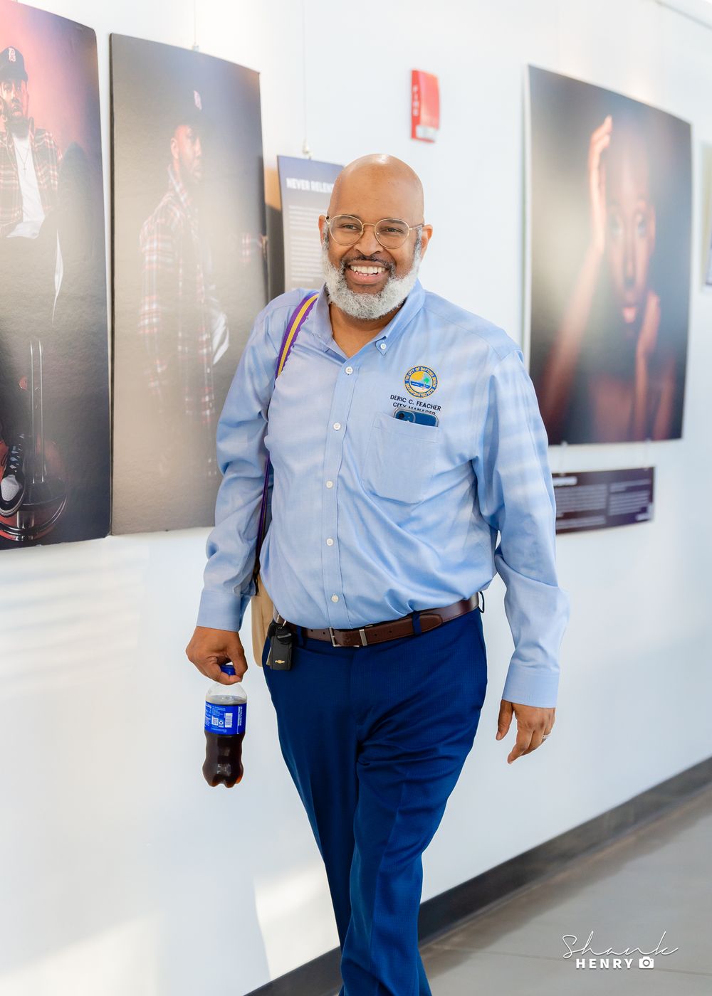 Professional in light blue button-down shirt and navy pants walking down hallway with framed artwork on wall.
