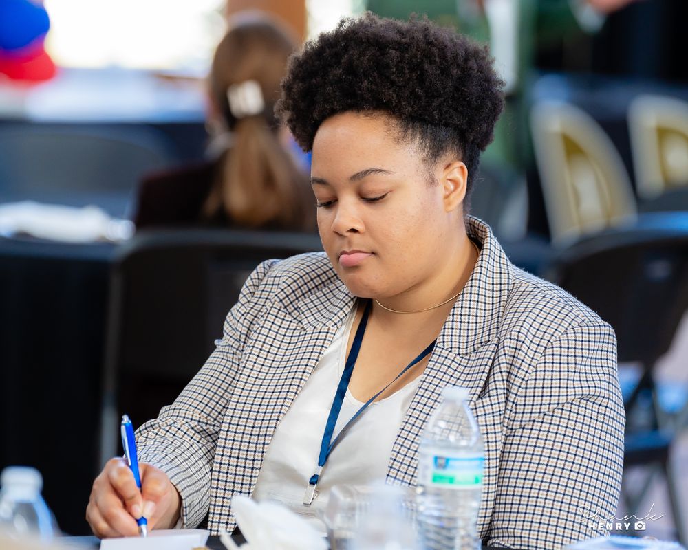 Person in checkered blazer writing notes during a meeting.