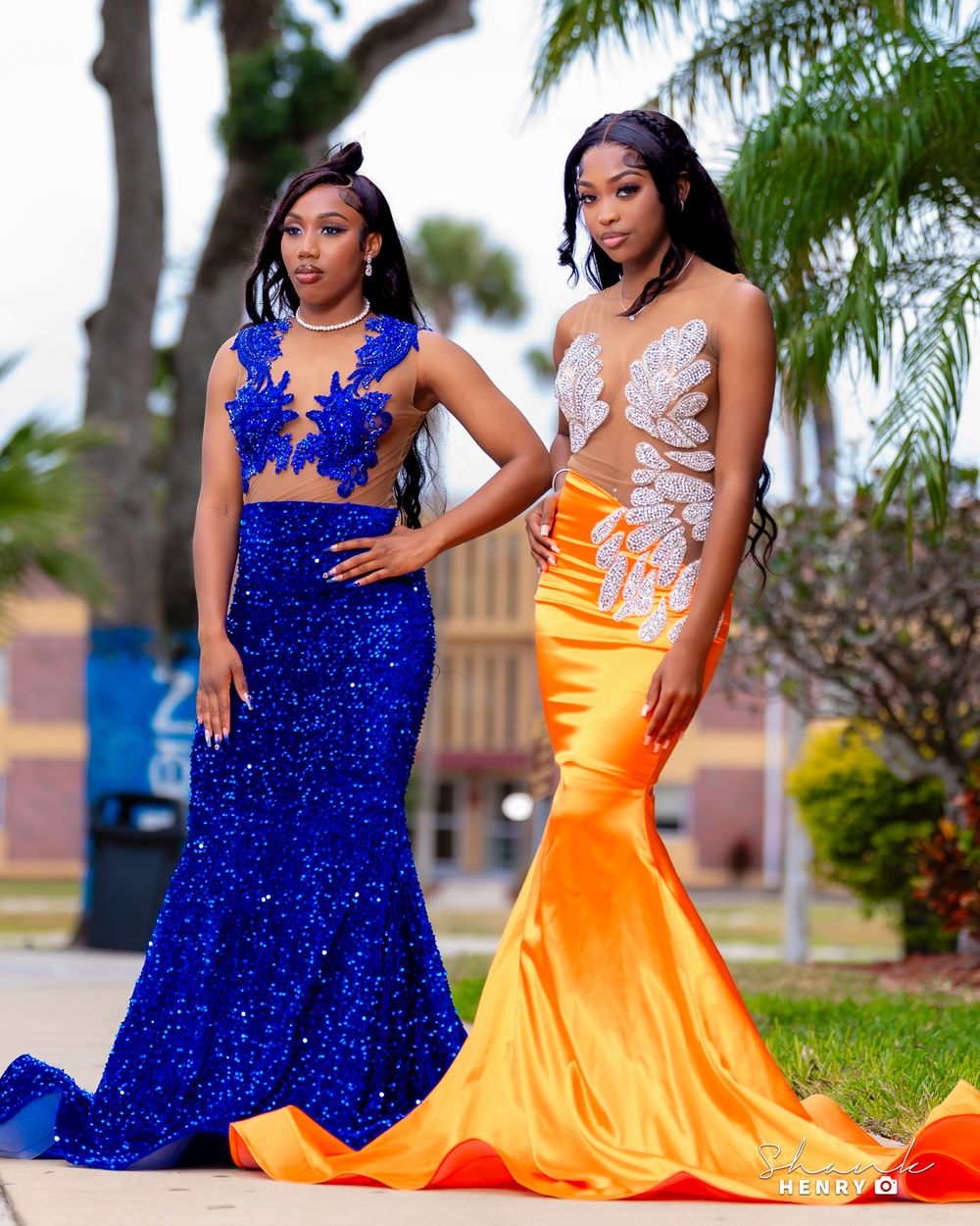Two women in formal gowns - one royal blue and one orange - pose outdoors.