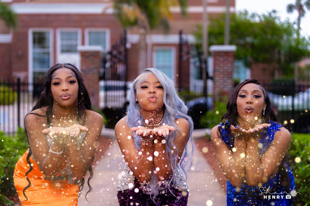 Three women in formal dresses throwing glitter in celebratory outdoor photo.