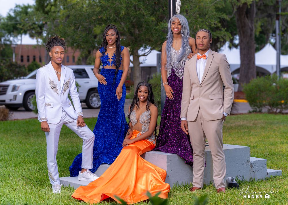 A group poses for a formal photo wearing elegant gowns and suits in white, blue, orange and purple.