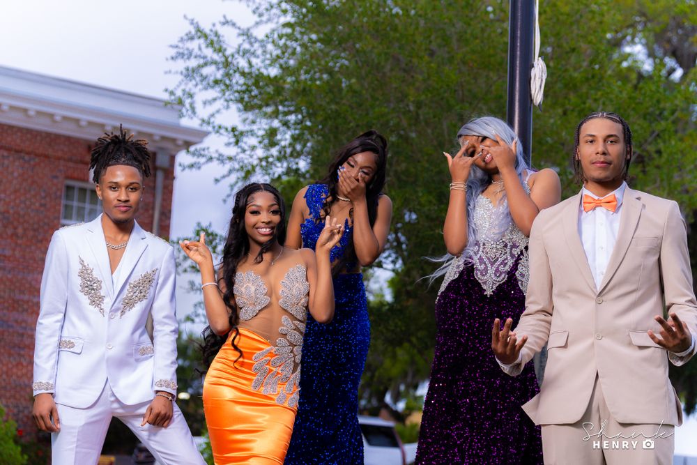 Friends celebrate in formal attire, striking playful poses outdoors near a brick building.