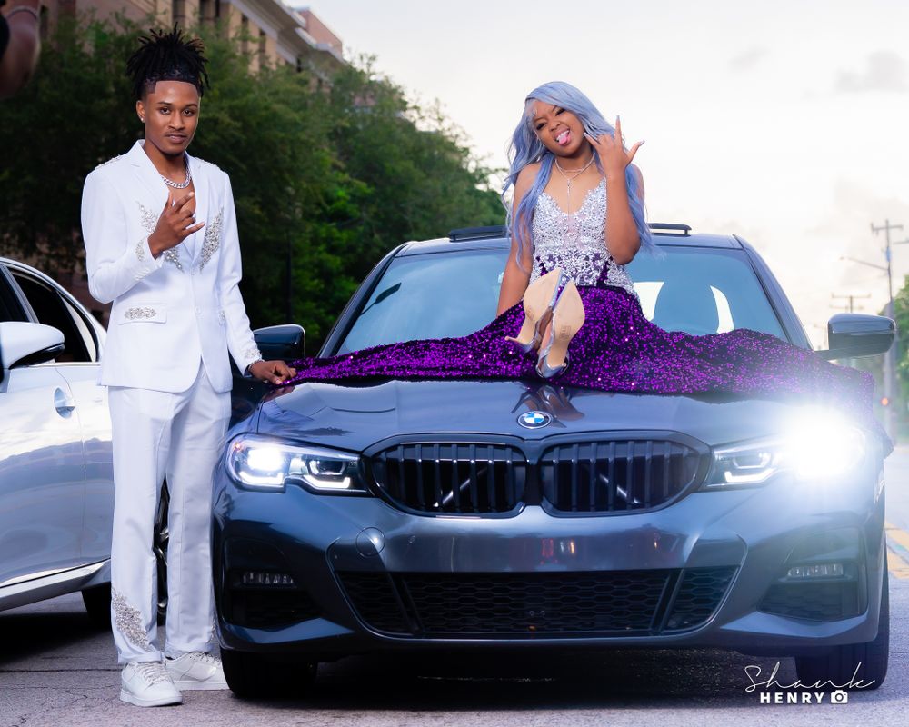 Two people pose with a sleek black BMW, one sitting on the hood wearing a sparkly purple dress.
