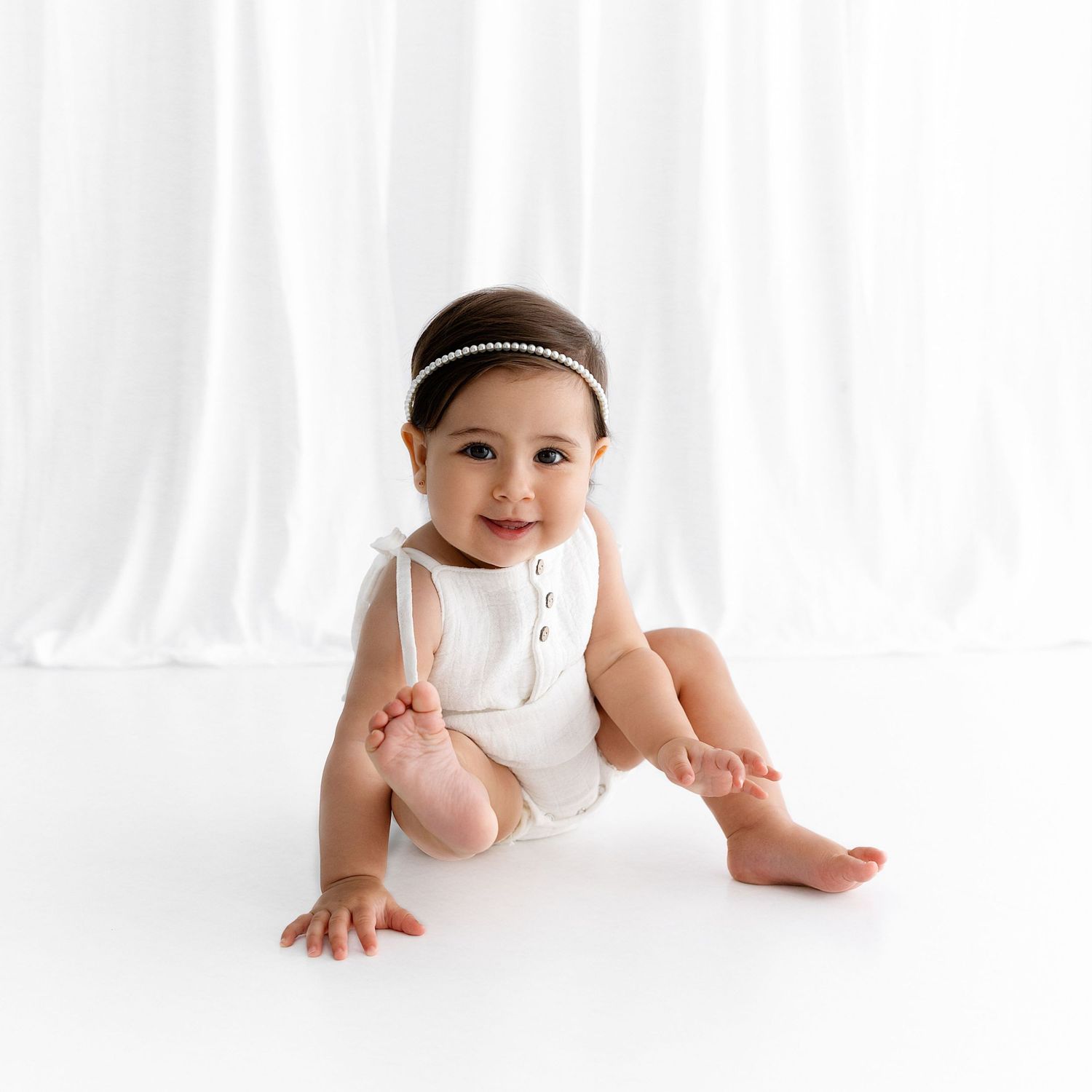 Baby in white romper and pearl headband sitting during photography session.