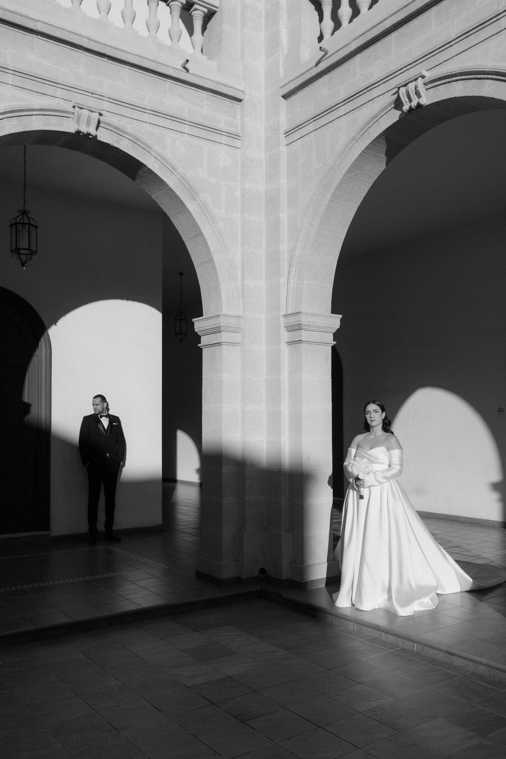 Black and white wedding photo showing a couple separated by elegant arched columns, with dramatic shadows on the walls.