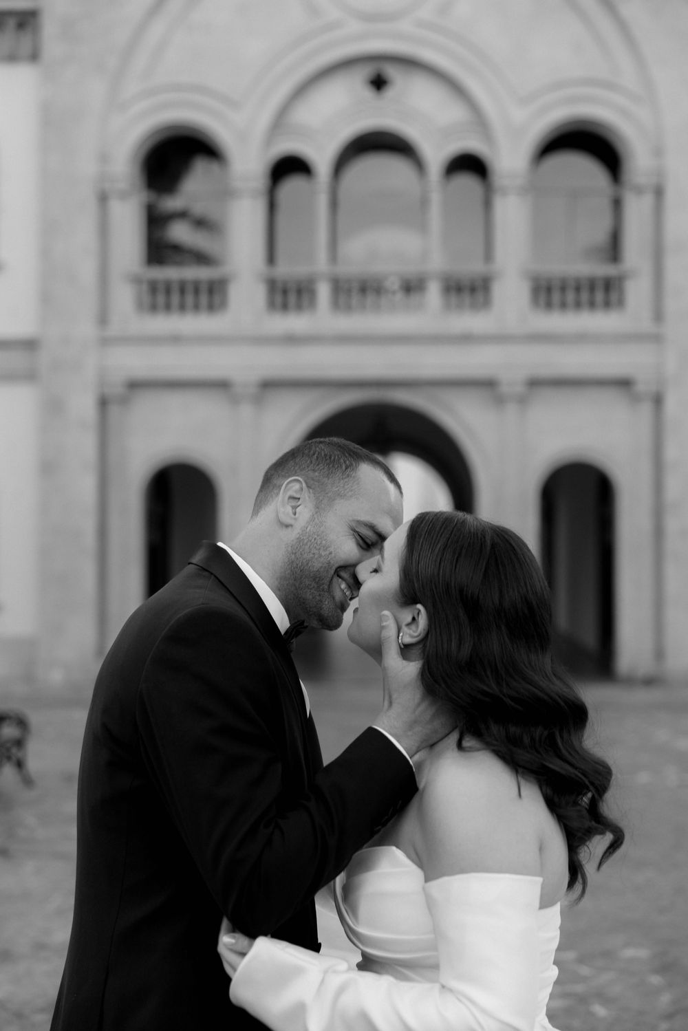 Black and white romantic wedding portrait showing a couple embracing and laughing in front of an elegant historic building.