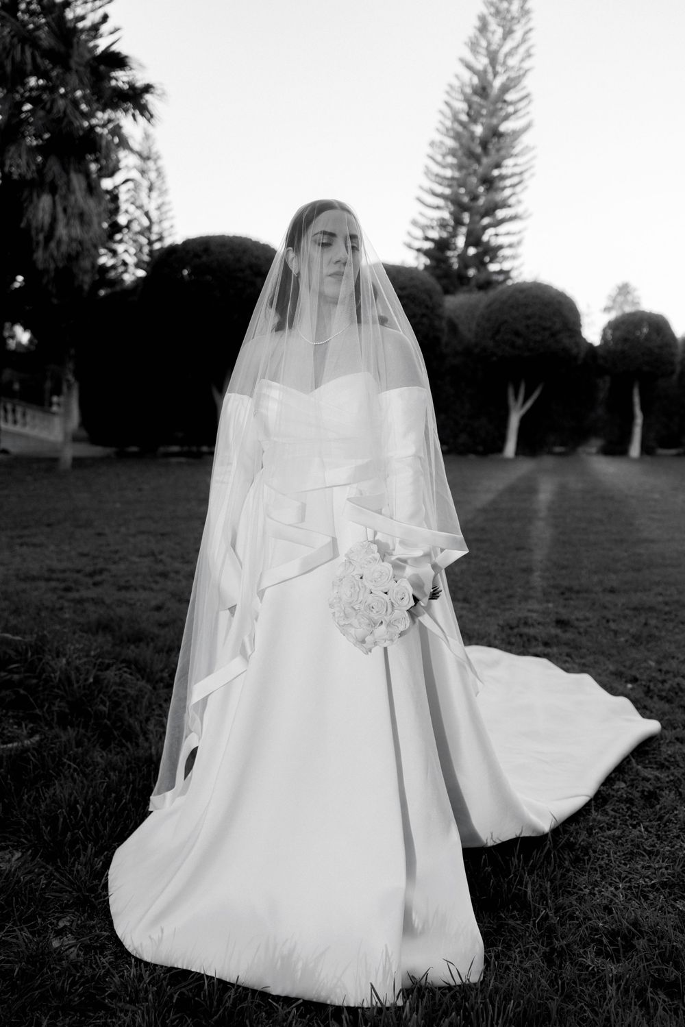 A black and white photograph of a bride in an elegant white wedding dress and long veil standing on garden grounds.