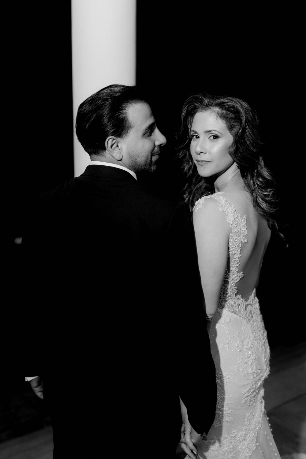 Elegant black and white portrait of a couple in formal attire, with bride in backless lace gown looking over her shoulder.