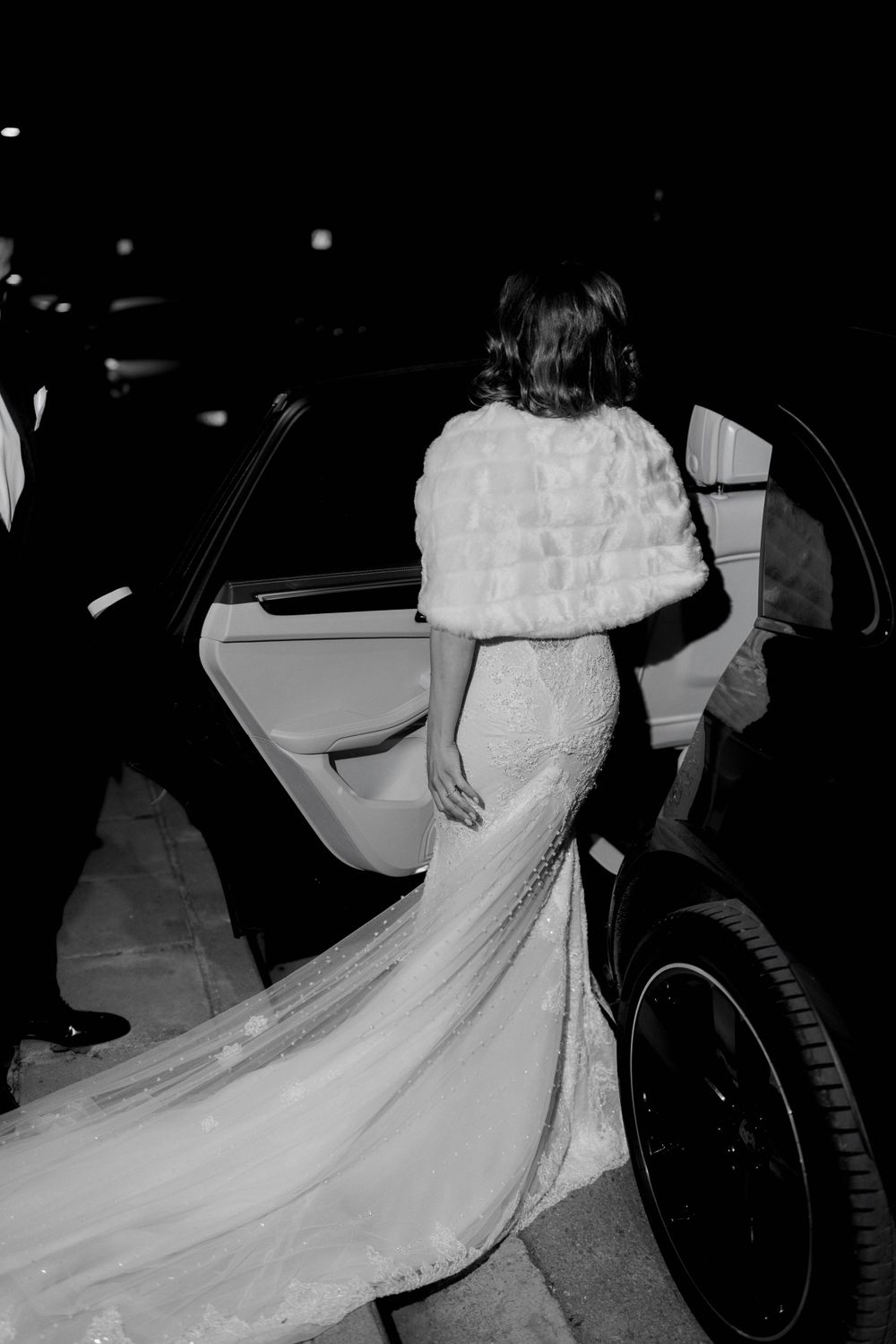 A bride in a white lace mermaid wedding gown with fur shawl steps into an elegant car at night.