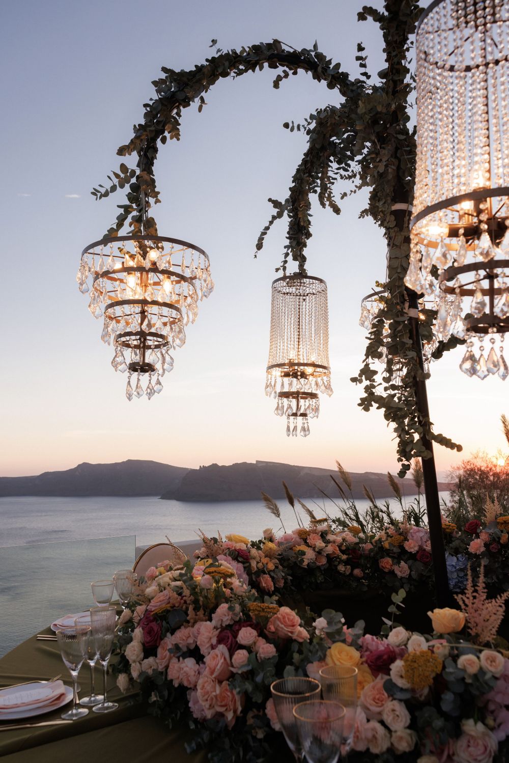Crystal chandeliers and floral arrangements decorate an outdoor wedding reception overlooking a sunset sea view.