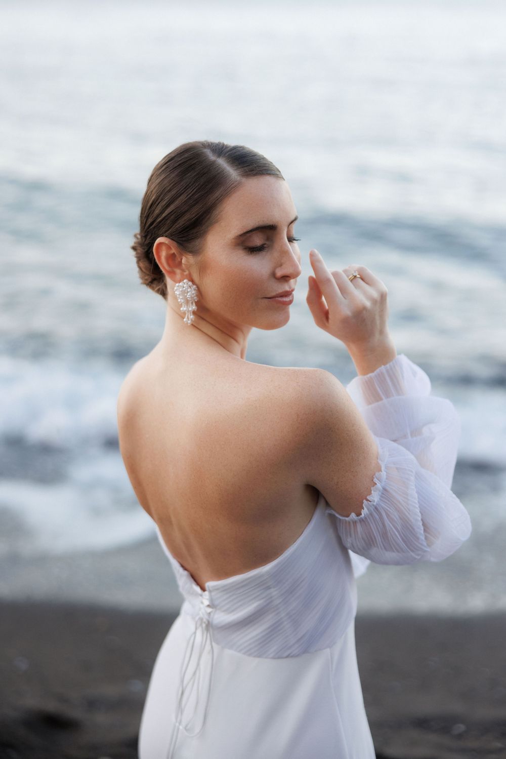 Artistic portrait of a woman in an elegant white off-shoulder dress posing on a moody beach at dusk.