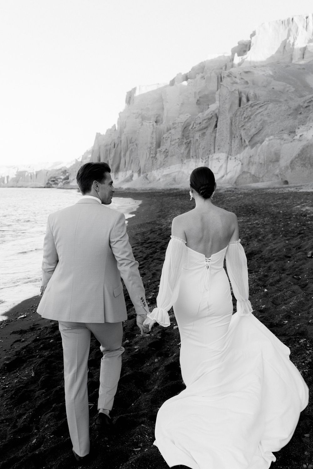 Newlywed couple walking on black sand beach with dramatic coastal cliffs in black and white photography.