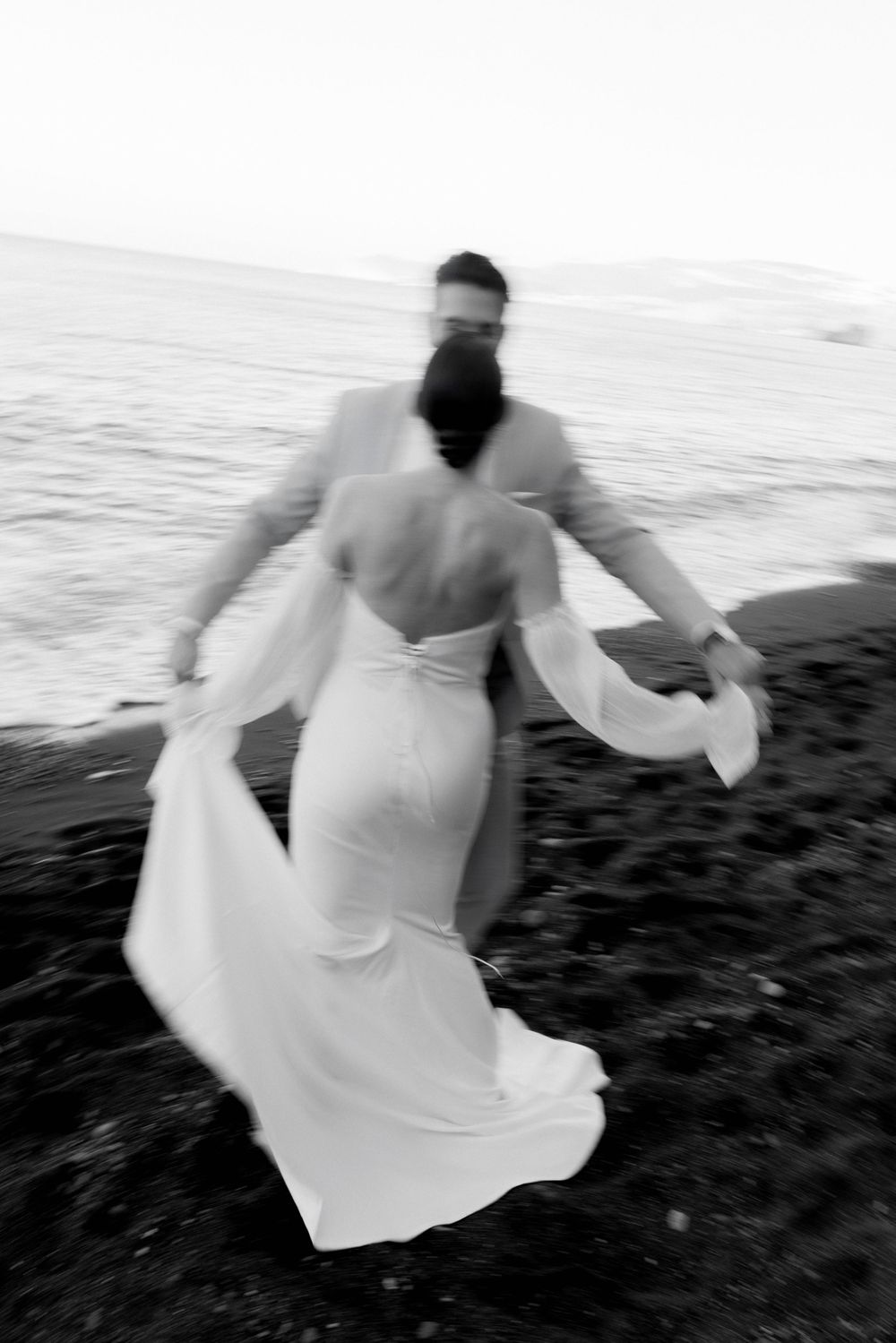 Black and white artistic photograph of a couple in wedding attire dancing on a beach with flowing fabric movement.