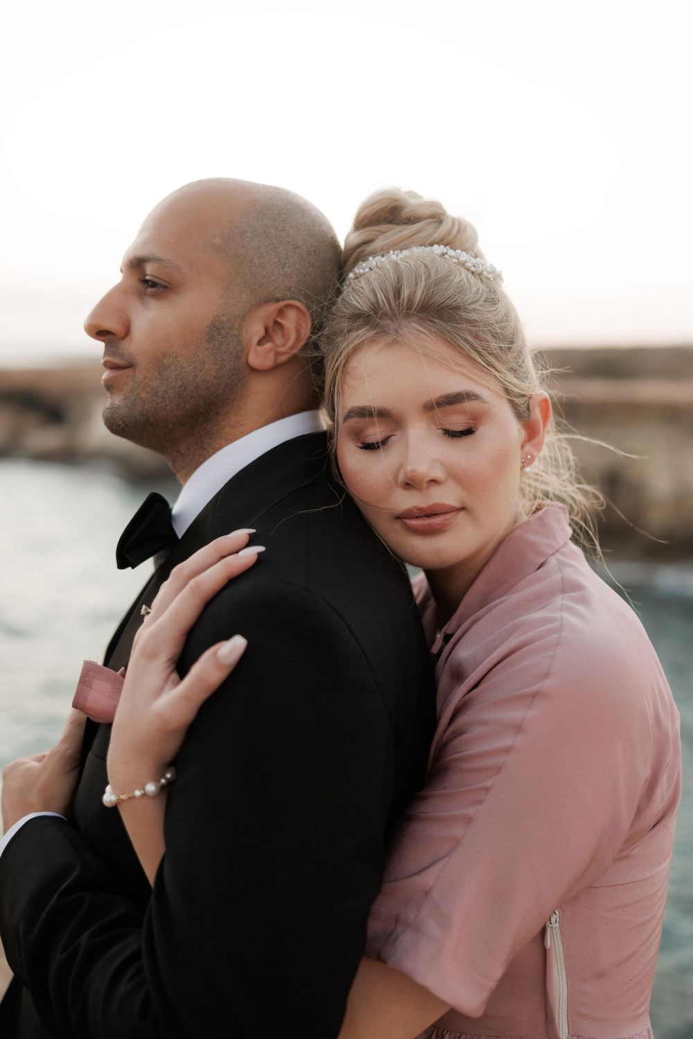Romantic couple poses together at sunset, with woman in pink embracing partner in black tuxedo from behind.