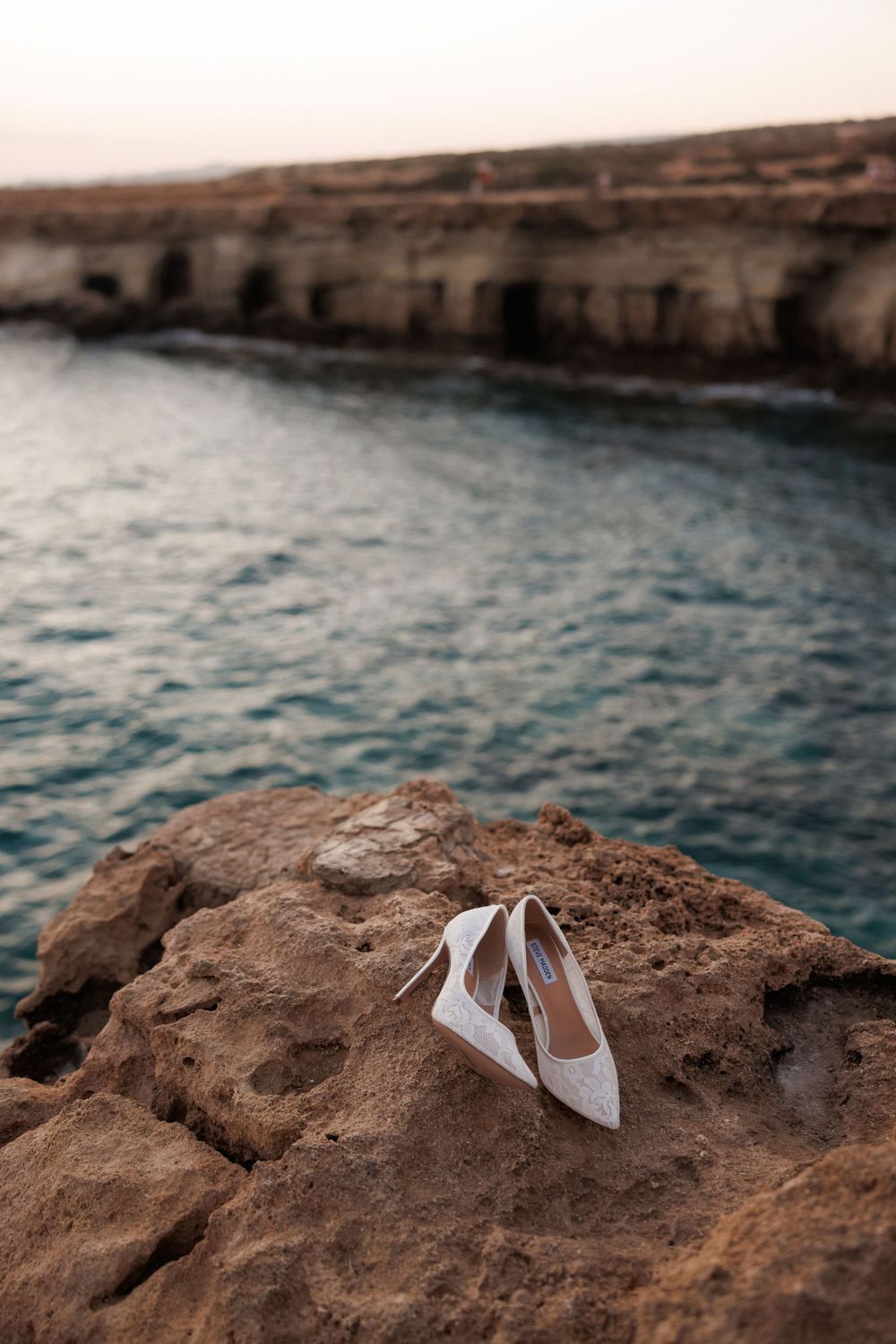 A pair of elegant white bridal heels positioned on rocky coastal cliffs overlooking blue ocean waters at sunset.