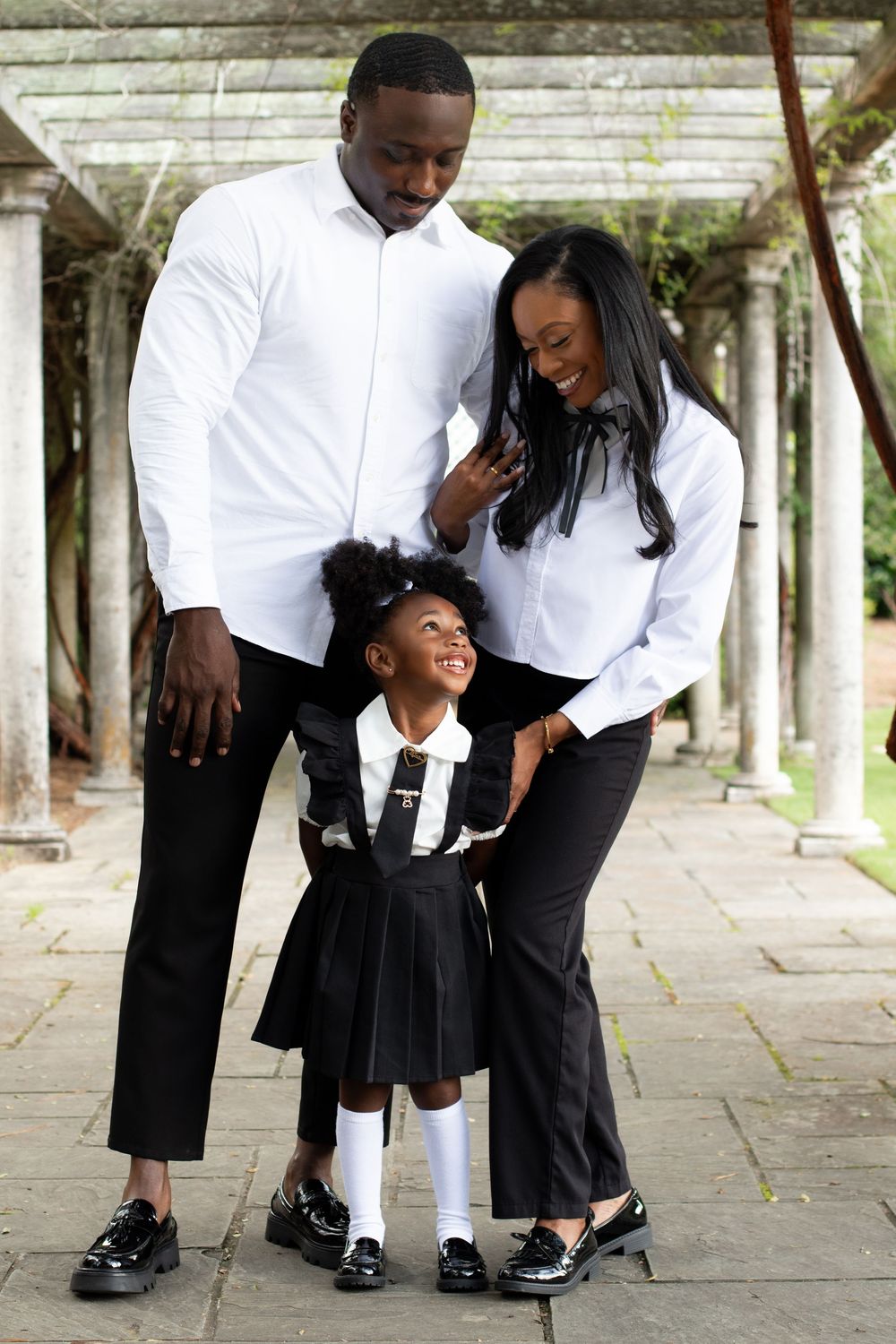 A happy Black family of three poses outdoors, parents in white shirts and black pants with their smiling daughter in a school uniform.