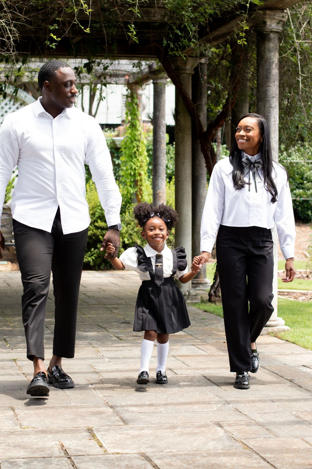 Happy Black family walking together outdoors, parents in black and white outfits with young daughter in school uniform.