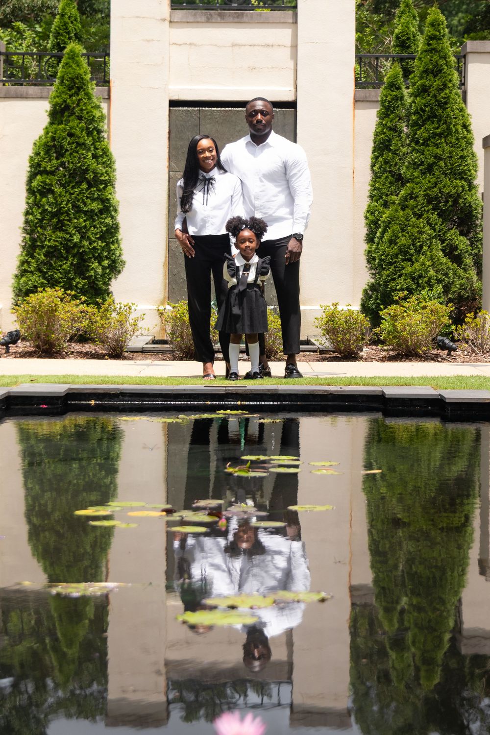 Family of three posing by a reflective pond in a lush garden with tall cypress trees.