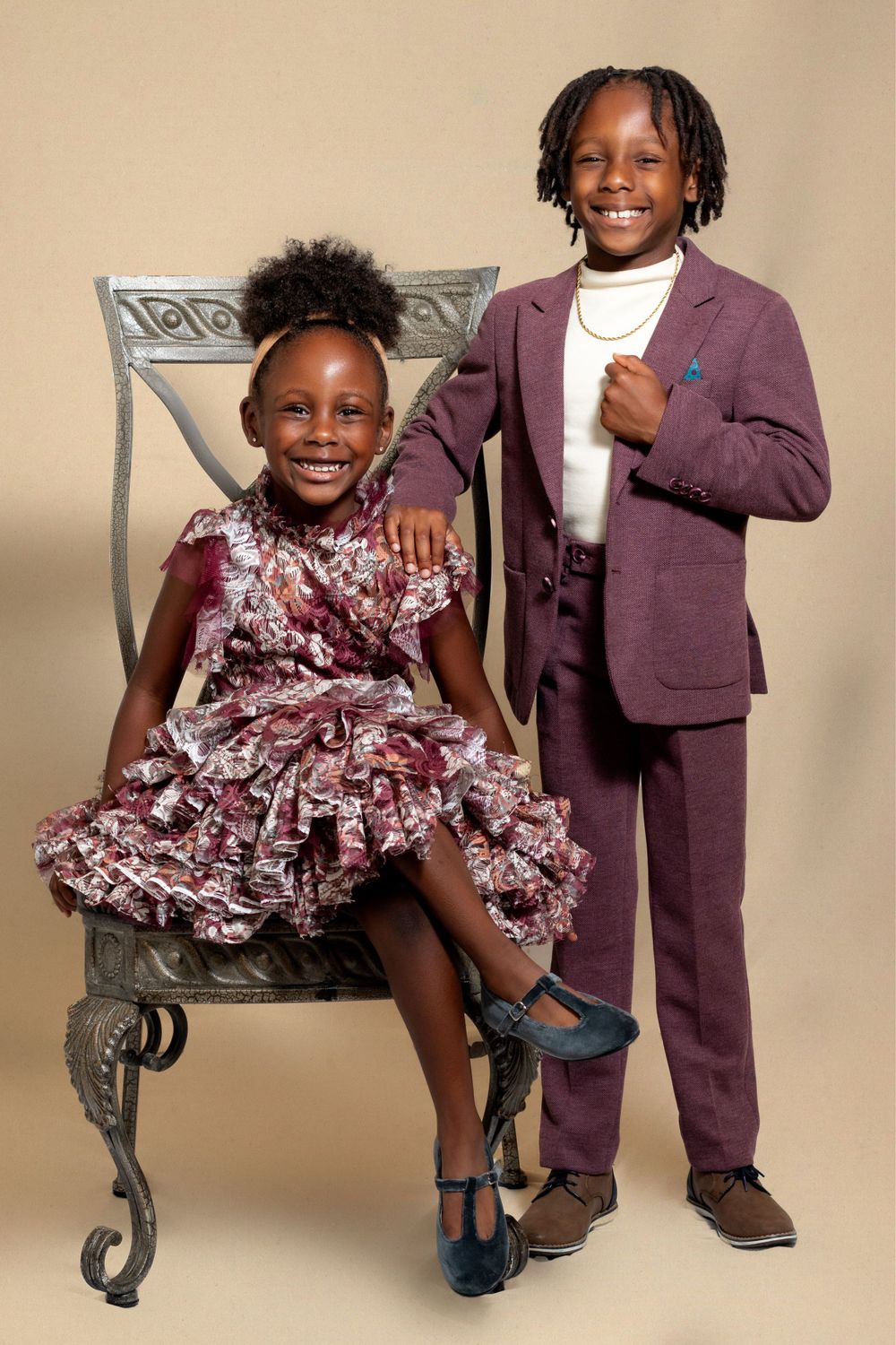 Two children dressed in formal attire pose in studio portrait against beige backdrop with ornate chair.